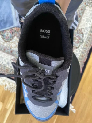 Gråa och blå sneakers från BOSS - Snygga sneakers från BOSS med grå ovandel, svarta detaljer och blå inslag. Skon har Ortholite-innersula för extra komfort och en robust, svartvit sula med tydliga ränder undertill. Materialmix av textil och syntet ger en modern look. Perfekt för dig som gillar stilrena och sportiga skor. Har också ett par till