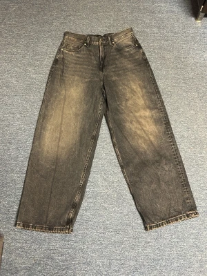 Baggy svarta jeans med tvättad look - Säljer ett par baggy svarta jeans med tvättad, lite blekt finish och raka ben. Jeansen har klassisk femficksmodell och hög midja. Perfekta för dig som gillar en avslappnad och trendig stil. Materialet är kraftig denim i bomull.
