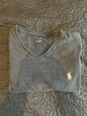 Mörkgrå v-ringad t-shirt från Ralph Lauren - Snygg mörkgrå t-shirt från Ralph Lauren med klassisk v-ringning och den ikoniska broderade loggan på bröstet. Tillverkad i mjuk bomull som känns skön mot huden. Perfekt till jeans eller shorts för en clean och enkel stil. Postas dagen eller dagen efter köpet.