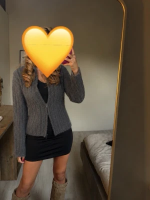 Grå stickad kofta med dragkedja - Mysig grå stickad kofta med dragkedja framtill och långa ärmar. Koftan har en rak passform och är perfekt att slänga över en klänning eller topp. Materialet ser ut att vara mjukt och värmande, perfekt för kyligare dagar.