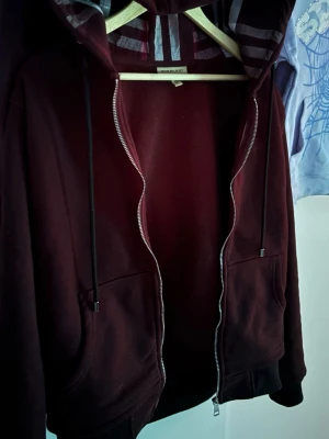 Burberry hoodie med dragkedja - Snygg hoodie från Burberry med dragkedja framtill och klassiska ränder i huvan. Finns i både vinröd och svart färg. Mjukt material, två fickor och justerbar huva med snören. Perfekt för en chill och stilren look.