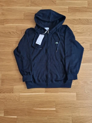 Svart hoodie från Lacoste - Snygg svart hoodie från Lacoste med dragkedja framtill och klassisk krokodillogga på bröstet. Tröjan har huva med dragsko, ribbade muddar och två fickor framtill. Perfekt för en avslappnad och sportig stil.