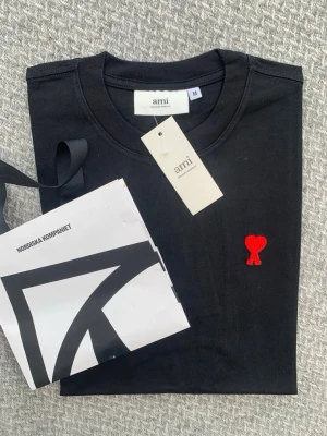 Svart t-shirt från Ami med rött hjärta - Snygg svart t-shirt från Ami Alexandre Mattiussi med klassisk passform och rund hals. På bröstet finns det ikoniska röda hjärta-loggan broderad. Perfekt för dig som gillar stilrena och trendiga plagg. Materialet är mjukt och bekvämt bomullstyg.