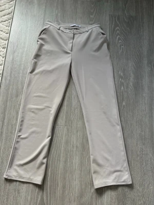 Beige kostymbyxor med raka ben - Snygga beige kostymbyxor med raka ben och klassisk passform. Byxorna har hällor för skärp, fickor fram och stängs med knapp och dragkedja. Perfekta för en stilren look. Märke syns ej, material okänt men känns mjukt och följsamt.
