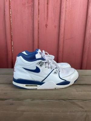 Nike vita och blå high sneakers - Nike sneakers i high-modell med vit ovandel, blå detaljer och röd logga på sulan. Skorna har klassisk snörning, vadderad krage och synlig Air-enhet i sulan. Materialet är mestadels läder med inslag av syntet. Perfekta för dig som gillar retrostil och sportig vibe.
