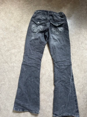 Grå bootcut jeans med broderi - Säljer ett par grå bootcut jeans med snygga broderade detaljer på bakfickorna och knappar. Jeansen har resår i midjan och klassisk femficksmodell. Perfekta för dig som gillar en avslappnad men ändå unik stil.