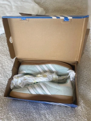 Adidas Gazelle mintgröna sneakers - Fräscha Adidas Gazelle sneakers i mintgrönt mocka med klassiska vita ränder och gummisula. Snygg retrostil med låg profil och diskret gulddetalj på sidan. Perfekta för dig som gillar streetwear och vill sticka ut med en unik färg.
