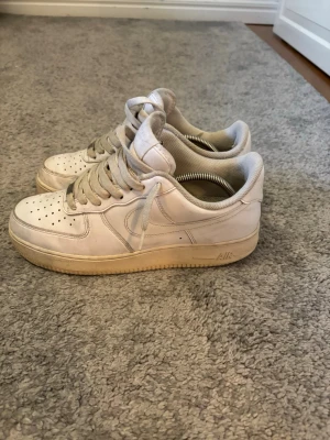 Vita Nike Air Force 1 sneakers - Klassiska vita Nike Air Force 1 sneakers med låg profil och perforerad tå för ventilation. Skorna har en tjock sula med AIR-dämpning och ikonisk Swoosh-logga på sidan. Tillverkade i läder med textildetaljer och vita skosnören.