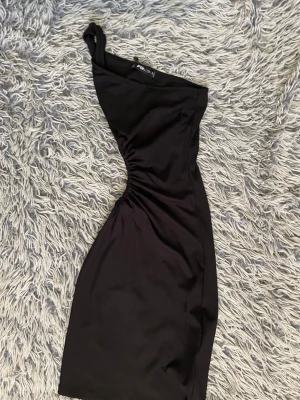 Svart oneshoulder klänning från RFS - Snygg svart oneshoulder klänning från RFS med tight passform och rynkade detaljer i midjan. Klänningen är kort och har en axelbandslös design på ena sidan, vilket ger en trendig look. Perfekt för dig som vill sticka ut.