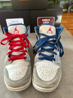 Air Jordan 1 Mid blå/vit - Säljer ett par Air Jordan 1 Mid sneakers i vitt och ljusblått med grå Swoosh och röda detaljer. Ena skon har röda skosnören och den andra blå. Klassisk Jumpman-logga på plösen och Air Jordan Wings på sidan. Ovandelen är i läder och textil.