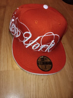 Orange New Era 59FIFTY keps New York - Snygg orange keps från New Era, modell 59FIFTY, med platt skärm och broderad vit text 'New York' i stor stil över framsidan. Kepsen har klassisk passform och sticker ut med sin färg och design. Perfekt för dig som vill ha en statement-accessoar.