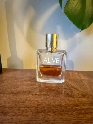 Hugo Boss Alive parfym  - Säljer en stilren parfymflaska från Hugo Boss med namnet ALIVE. Flaskan är rektangulär i genomskinligt glas med guldig metallkork och vit text. Innehållet är bärnstensfärgat. Perfekt för dig som gillar eleganta och moderna dofter.