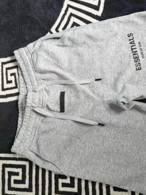 Grå Essentials shorts med snörning - Snygga grå shorts från Essentials Fear of God med elastisk midja och snörning framtill. Mjukt material och logga tryckt på ena benet. Perfekta för en chill och avslappnad stil.