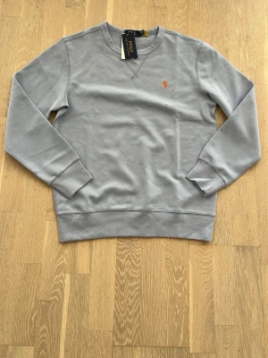 Grå sweatshirt från Polo Ralph Lauren - Snygg grå sweatshirt från Polo Ralph Lauren med klassisk rund halsringning och ribbade muddar. Tröjan har den ikoniska orangea logotypen broderad på bröstet. Perfekt för en avslappnad och stilren look.