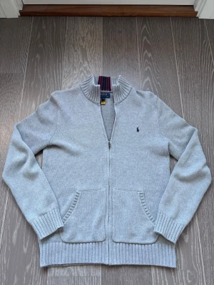 Polo Ralph Lauren helzip - Snygg grå ziptröja från Polo Ralph Lauren med ribbad krage och muddar. Tröjan har två fickor framtill och en diskret logga broderad på bröstet. Passar som storlek S. Hör av er vid frågor!