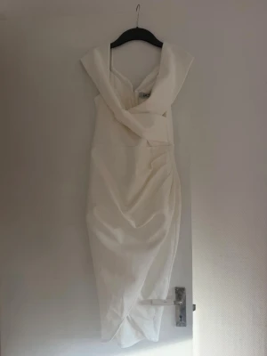 Vit omlottklänning med drapering - Elegant vit långklänning med omlott-detalj och draperad front. Klänningen har breda axelband och en v-ringad urringning som ger en stilren och modern look. Perfekt för dig som vill sticka ut med en sofistikerad stil.