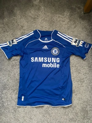 Chelsea matchtröja Drogba #11 Adidas - Blå Chelsea matchtröja från Adidas med Drogba och nummer 11 på ryggen. Tröjan har vita detaljer, Premier League-patch på ärmarna och Samsung Mobile som sponsortryck. Tillverkad i lätt och ventilerande material, perfekt för fotboll eller samling.