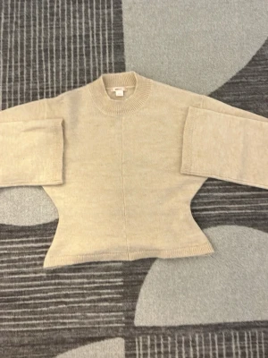 Beige stickad tröja från Gina Tricot - Snygg beige stickad tröja från Gina Tricot i storlek XS. Tröjan har breda ärmar, markerad midja och ribbstickad halsringning. Perfekt för dig som gillar en enkel men trendig look. Materialet känns mjukt och skönt mot huden.