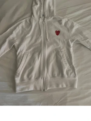 Vit hoodie från Comme des Garçons - Snygg vit hoodie med dragkedja från Comme des Garçons Play. Har huva med dragsko, två fickor fram och det ikoniska röda hjärtloggan på bröstet. Perfekt för en clean och stilren look.