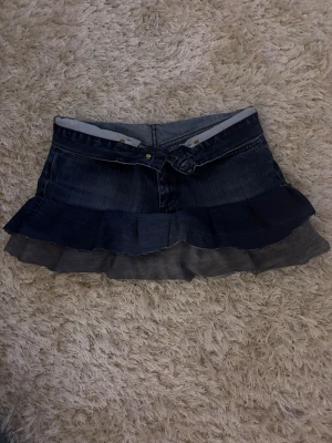 Mörkblå volangkjol i denim - Snygg kort jeanskjol i mörkblå denim med volangdetaljer i två lager. Kjolen har klassisk femficksdesign och knäppning framtill. Perfekt för dig som gillar en trendig och ungdomlig stil.  Kjolen är köpt på Plick men används inte då den är lite för stor  . 