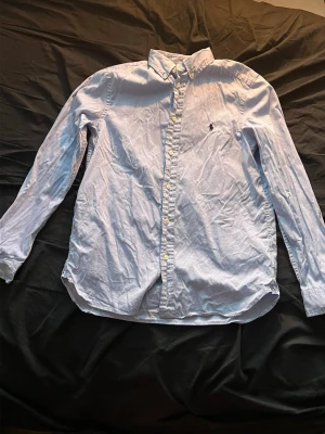 Blå skjorta från Polo Ralph Lauren - Klassisk ljusblå skjorta från Polo Ralph Lauren i slim fit-modell. Skjortan har långa ärmar, knappar framtill och den ikoniska broderade loggan på bröstet. Tillverkad i mjuk bomull och passar perfekt till en stilren look.