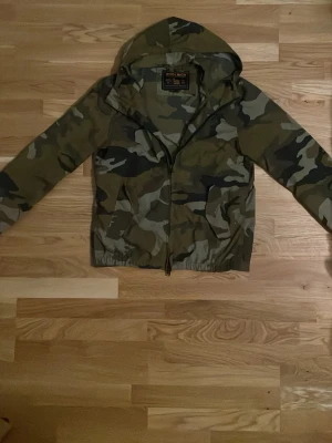 Camo Woolrich Windbreaker  - Vi säljer den här stilrena windbreakern från Woolrich i en camo grön färg. Den är i väldigt bra skick utan stora täcken på användning och är i storlek S. Perfekt för sommaren och sommarkvällar. Hör av dig om du har frågor.