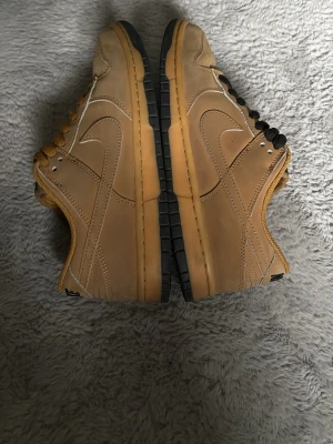 Nike Dunk Low beige och svart - Säljer ett par Nike Dunk Low sneakers i beige läder med svarta och senapsgula skosnören. Skorna har klassisk låg siluett, svart yttersula och tydliga sömmar längs sidorna. Perfekta för dig som gillar streetwear och vill sticka ut med snygga färgdetaljer. Kommer med både färgerna av knytsnören 