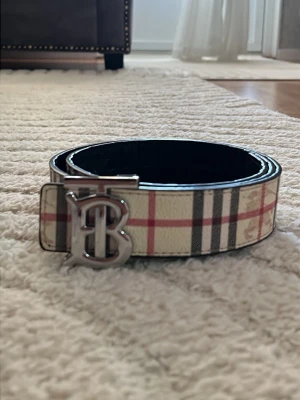 Burberry bälte med klassiskt rutmönster - Snyggt Burberry bälte med det ikoniska rutmönstret i beige, rött och svart. Bältet har ett stilrent metallspänne med Burberrys logotyp. Perfekt accessoar för att lyfta din outfit.