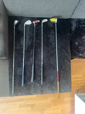 Golfklubbor mixade märken 5-pack - Fem golfklubbor i metall med olika greppfärger och klubbhuvuden. Märken som Silver, Cobra och Young 3.0 syns på skaften. Greppen är svarta, blå, grå och röda. Klubbhuvudena är silverfärgade, en med gul detalj. Perfekt för dig som vill ha ett varierat golfset.