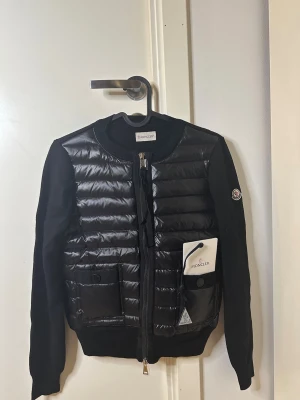 Svart cardiganjacka från Moncler - Snygg svart cardiganjacka från Moncler med quiltad, glansig front och ärmar i mjukt tyg. Rund halsringning, dragkedja framtill och fickor med lock. Moncler-logga på ärmen och ribbade muddar vid ärmslut och nederkant.