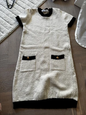 Beige kort klänning med svarta detaljer - Stilren beige kort klänning med svarta kanter vid ärmar, hals och nederkant. Två fickor framtill med svarta lock och guldfärgade knappar. Rak passform och kortärmad, perfekt för en trendig look.