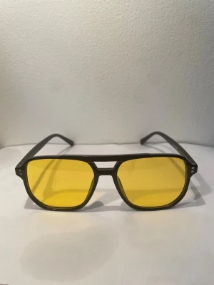 Gula solglasögon  - Coola solglasögon med svart båge och stora, fyrkantiga gula glas. Perfekt accessoar för dig som vill sticka ut och ge din stil en retrovibe. Bågarna är i plast och glasen har en tydlig gul ton.