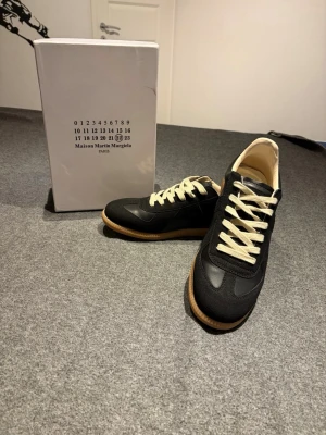 Maison Margiela svarta sneakers  - Svarta sneakers från Maison Martin Margiela med beigea skosnören och klassisk gummisula. Skorna har en stilren design i läder och mocka, med lågt skaft och diskreta detaljer. Perfekta för dig som gillar minimalistisk och trendig stil.