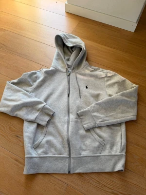 Grå zip-hoodie från Polo Ralph Lauren - Snygg ljusgrå hoodie med dragkedja från Polo Ralph Lauren. Klassisk design med huva, dragsko och två fickor framtill. Liten broderad logga på bröstet. Mjuk bomullsblandning som är skön att ha på sig.