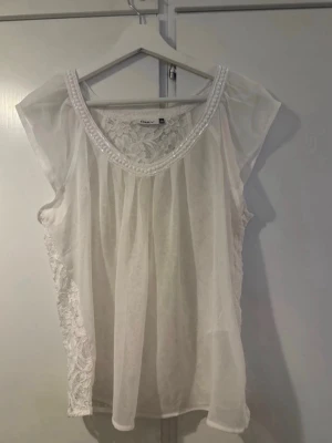 Vit blus med spetsdetaljer från Only - Söt vit blus från Only med transparenta partier och vackra spetsdetaljer på ryggen och sidorna. Blusen har rund halsringning med pärlbroderi och är ärmlös med lätt, luftig känsla. Perfekt för en fräsch och feminin look.