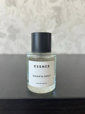 ESSNCE Warm'n Spicy Eau de Parfum - ESSNCE Warm'n Spicy Eau de Parfum i en stilren, rund glasflaska med svart lock. Doften har en varm och kryddig karaktär, perfekt för dig som gillar intensiva och unika parfymer. Flaskan har en minimalistisk vit etikett med svart text. Säljer då jag har för många parfymer
