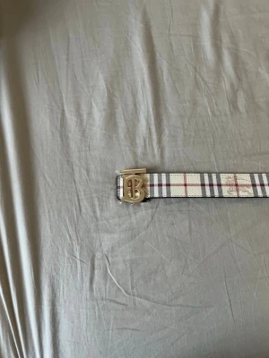 Burberry vändbart bälte med TB-spänne - Snyggt vändbart bälte från Burberry med klassiskt rutigt mönster i beige, svart och rött på ena sidan och svart på andra sidan. Bältet har ett guldigt TB-monogram som spänne och är gjort i skinn. Perfekt accessoar för att lyfta din outfit.