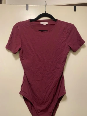 Vinröd kortärmad body från H&M - Säljer en vinröd body med korta ärmar och rund halsringning. Bodyn har en enkel och stilren design, perfekt att matcha med jeans eller kjol. Materialet känns mjukt och stretchigt för en bekväm passform. Bara använt en gång!