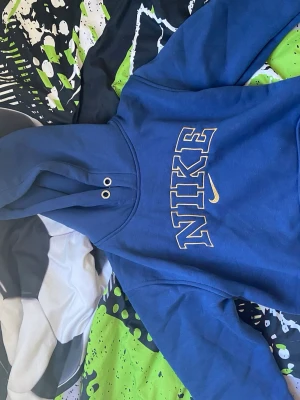 Blå hoodie från Nike med tryck - Blå hoodie från Nike med stort guldfärgat NIKE-tryck på bröstet och klassisk huva med metallöljetter. Mjuk insida och ribbade muddar. Perfekt för en avslappnad streetstyle-look.