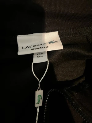 Svart hoodie från Lacoste, Regular Fit - Svart hoodie från Lacoste i regular fit med dragkedja framtill. Klassisk design med diskret Lacoste-logga och etikett. Perfekt för en stilren och avslappnad look. Materialet känns mjukt och bekvämt mot huden.