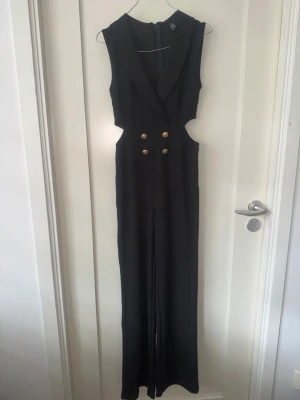 Svart ärmlös jumpsuit med cut-outs - Stilren svart ärmlös jumpsuit med djup v-ringning, markerad krage och snygga cut-outs i midjan. Framtill finns dekorativa guldknappar som ger en cool touch. Vida ben och dragkedja bak. Perfekt för dig som gillar trendiga och eleganta plagg.