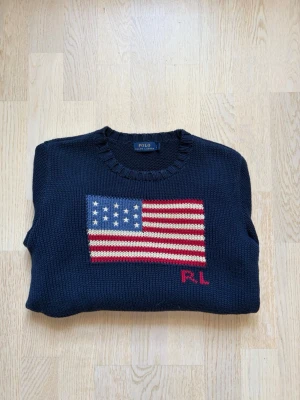Mörkblå stickad tröja Polo Ralph Lauren - Mörkblå stickad tröja från Polo Ralph Lauren med amerikansk flagga och RL-broderi på bröstet. Tröjan har rund halsringning och ribbstickade muddar vid ärmslut och nederkant. Säljer denna då den helt enkelt inte användes mer men är i ett riktigt fint skick, inga defekter, inga utstretchningar, som ny!!! Glöm inte att följare får rabatt, så…. följ! Bara att skriva för bilder, mått och sånt!
