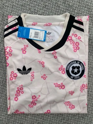 Vit Adidas fotbollströja Chile L - Vit fotbollströja från Adidas med svarta detaljer och rosa blommönster. Tröjan har svart V-ringad krage, tre svarta ränder på axlarna och patch med Federación de Fútbol de Chile på bröstet. Tillverkad i lätt och ventilerande material.