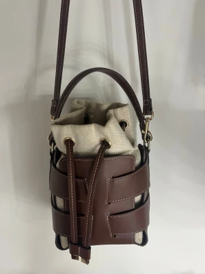 Brun bucket bag från H&M - Trendig bucket bag i brunt läder från H&M med justerbar axelrem och handtag. Väskan har ett unikt flätat mönster och en innerpåse i beige canvas med dragsko. Perfekt för dig som gillar stilrena och moderna accessoarer.