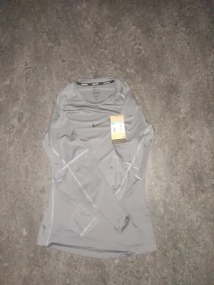 Grå långärmad träningströja Nike - Snygg grå långärmad träningströja från Nike med diskret logga på bröstet. Tillverkad i ett lätt och stretchigt funktionsmaterial som andas, perfekt för löpning eller gym. Tröjan har rund halsringning och smal passform.