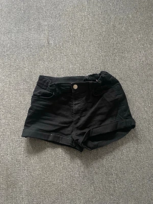 Svarta jeansshorts med upprullad kant - Snygga svarta jeansshorts med upprullad kant och klassisk femficksdesign. Stängs med knapp och dragkedja framtill. Perfekta för sommardagar och enkla att matcha med olika toppar. Materialet är denim vilket ger en cool och avslappnad look.