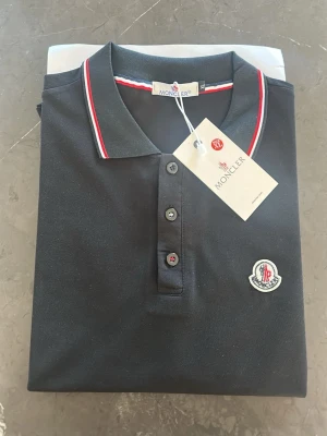 Svart Moncler polotopp med logga - Säljer en svart polotopp från Moncler med klassisk krage och knappar framtill. Toppens krage och ärmslut har snygga röda och vita detaljer. Moncler-loggan är broderad på bröstet. Perfekt för dig som gillar stilrena och exklusiva plagg. Storlek XL men passar även L 