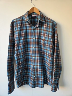 Peak Performance mens shirt size M - Snygg långärmad skjorta från peak Performance i blått, brunt och vitt rutmönster. Klassisk krage, bröstficka och knäppning framtill. Perfekt för dig som gillar en avslappnad men stilren look. Skjortan är i mjukt bomullstyg och har en tidlös passform. Size M