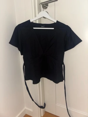Omlott top med knytning - Stilren svart omlottopp från Gina Tricot med korta ärmar och knytband i midjan. Toppen har v-ringning och en lätt figurnära passform som framhäver siluetten. Perfekt att matcha med jeans eller kjol för en trendig look.