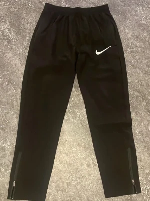 Svarta träningsbyxor från Nike - Säljer ett par svarta träningsbyxor från Nike med vit logga på låret. Byxorna har dragkedjor vid bensluten och resår i midjan. Perfekta för träning eller chill. Materialet är mjukt och stretchigt för bästa komfort.